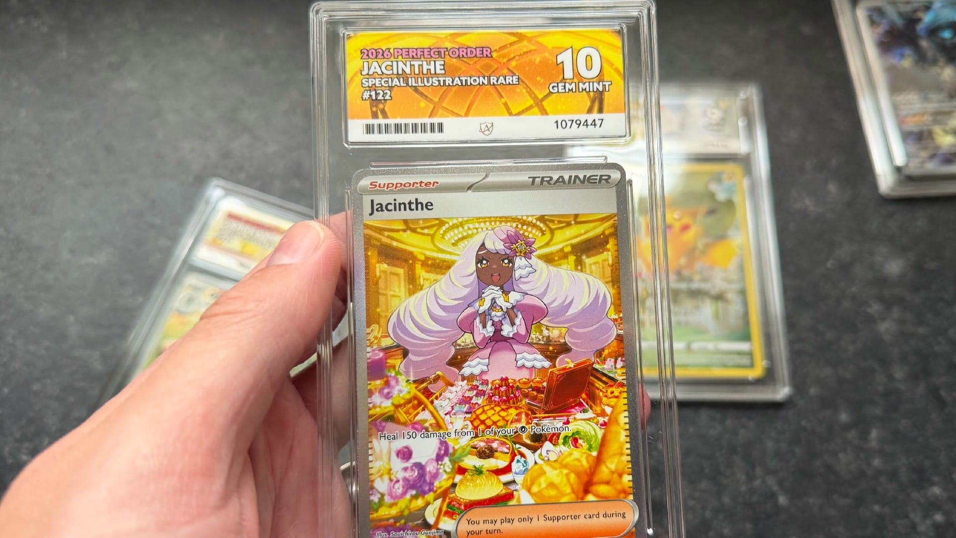 A hand holding a Jacinthe Pokemon card with an ACE 10 slab
